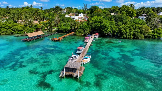 Vista aérea de un muelle y embarcaciones entre agua turquesa y vegetación.